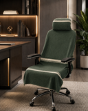 A dark green office chair with a matching headrest and seat cover draped over it, set in a modern home office environment with a desk, bookshelf, and potted plant.