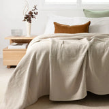 A beige waffle-weave throw blanket draped over a bed in a bright, modern bedroom setting with white pillows, a brown accent pillow, and a wooden bedside table.