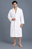 A man wearing a white cotton bathrobe with a shawl collar, waist tie, and front patch pockets, standing against a solid gray studio background.